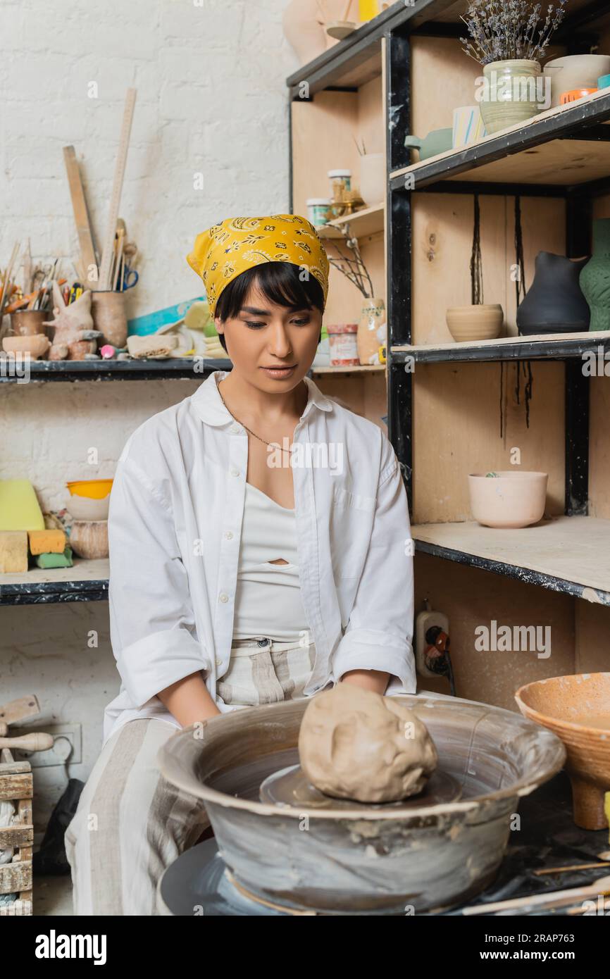 Young asian female artisan in headscarf and workwear looking at clay on