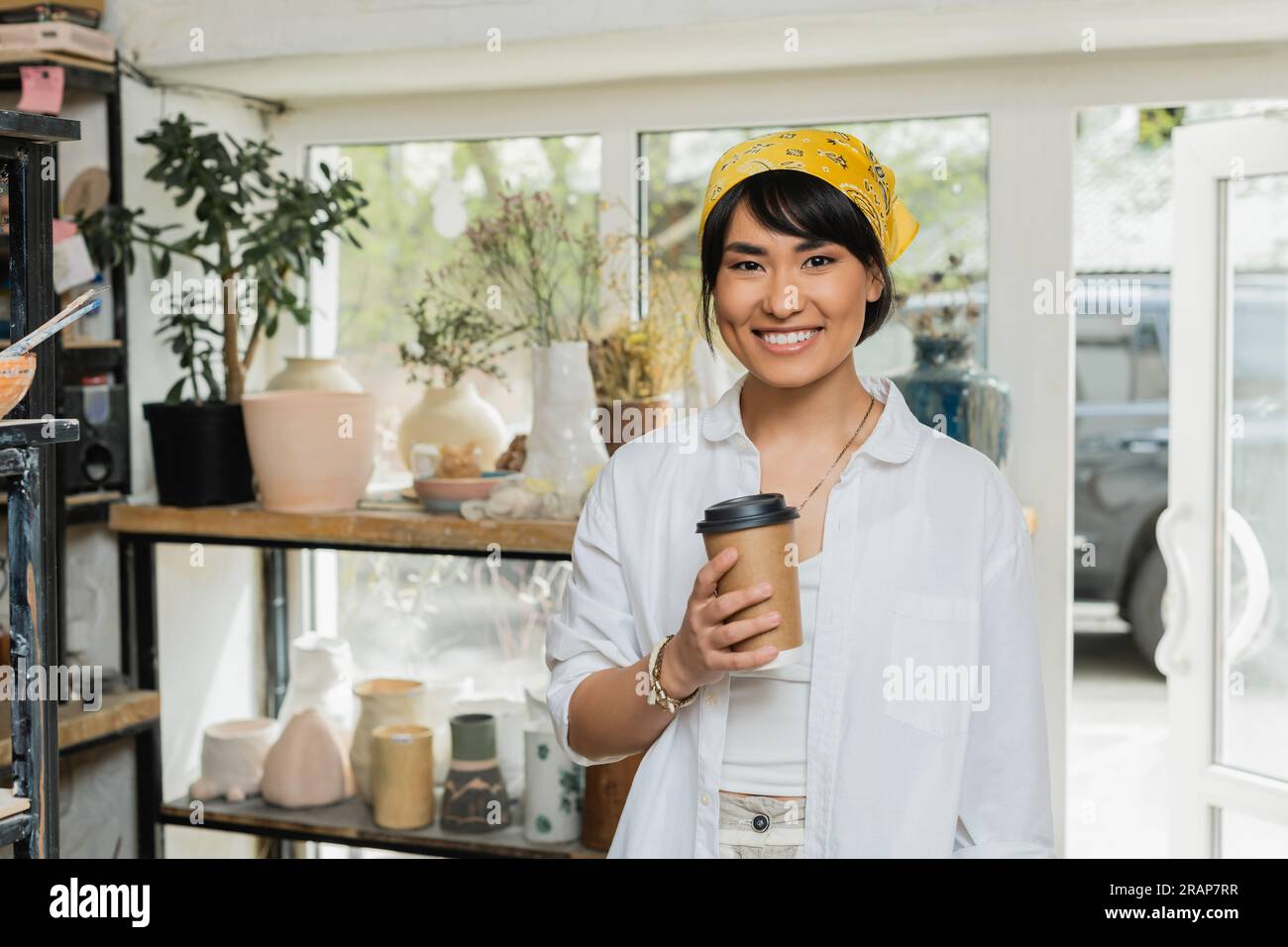 Positive young asian artist in headscarf and workwear looking at camera and holding coffee to go