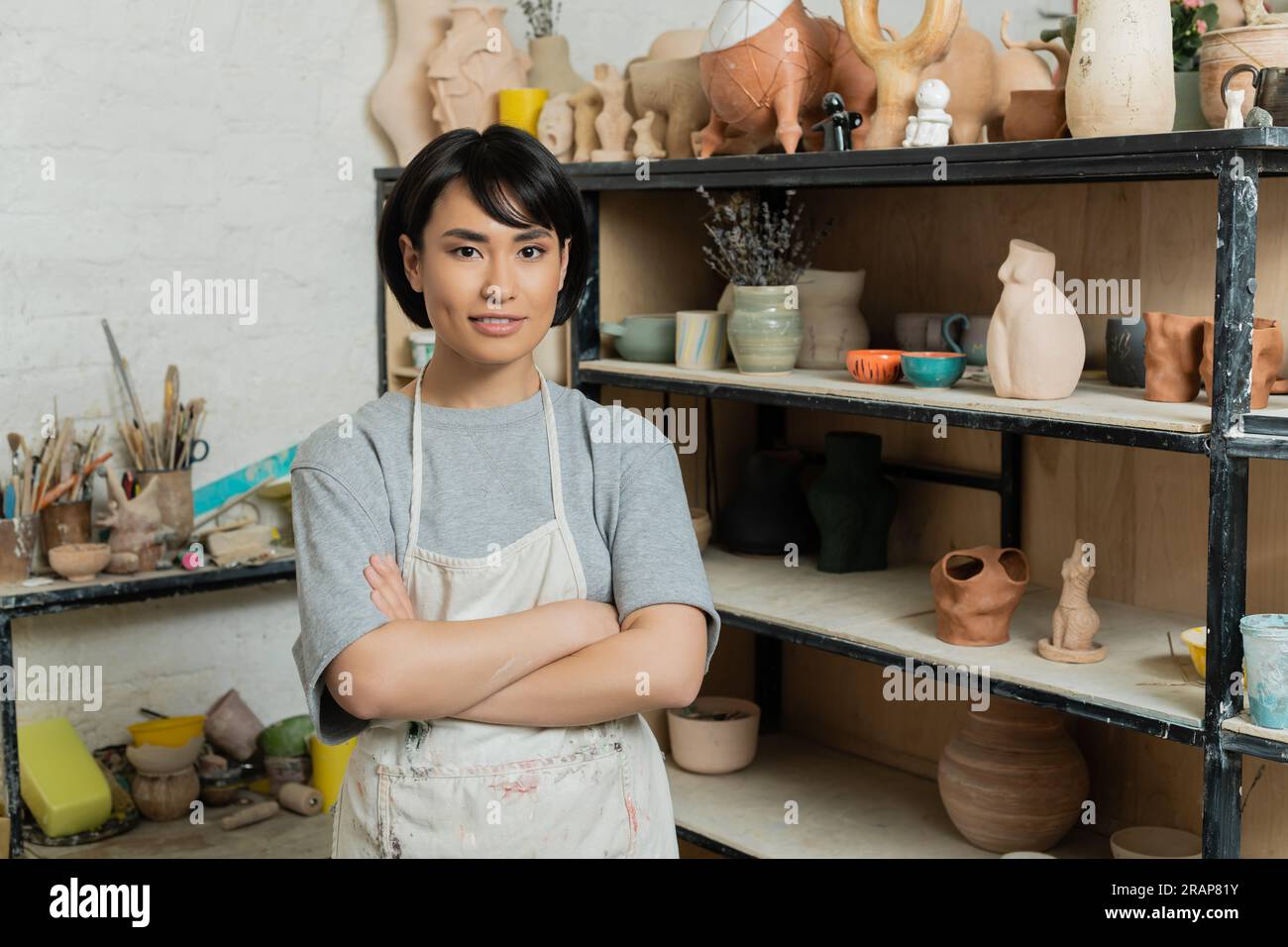 Smiling young asian female artisan in apron crossing arms and looking