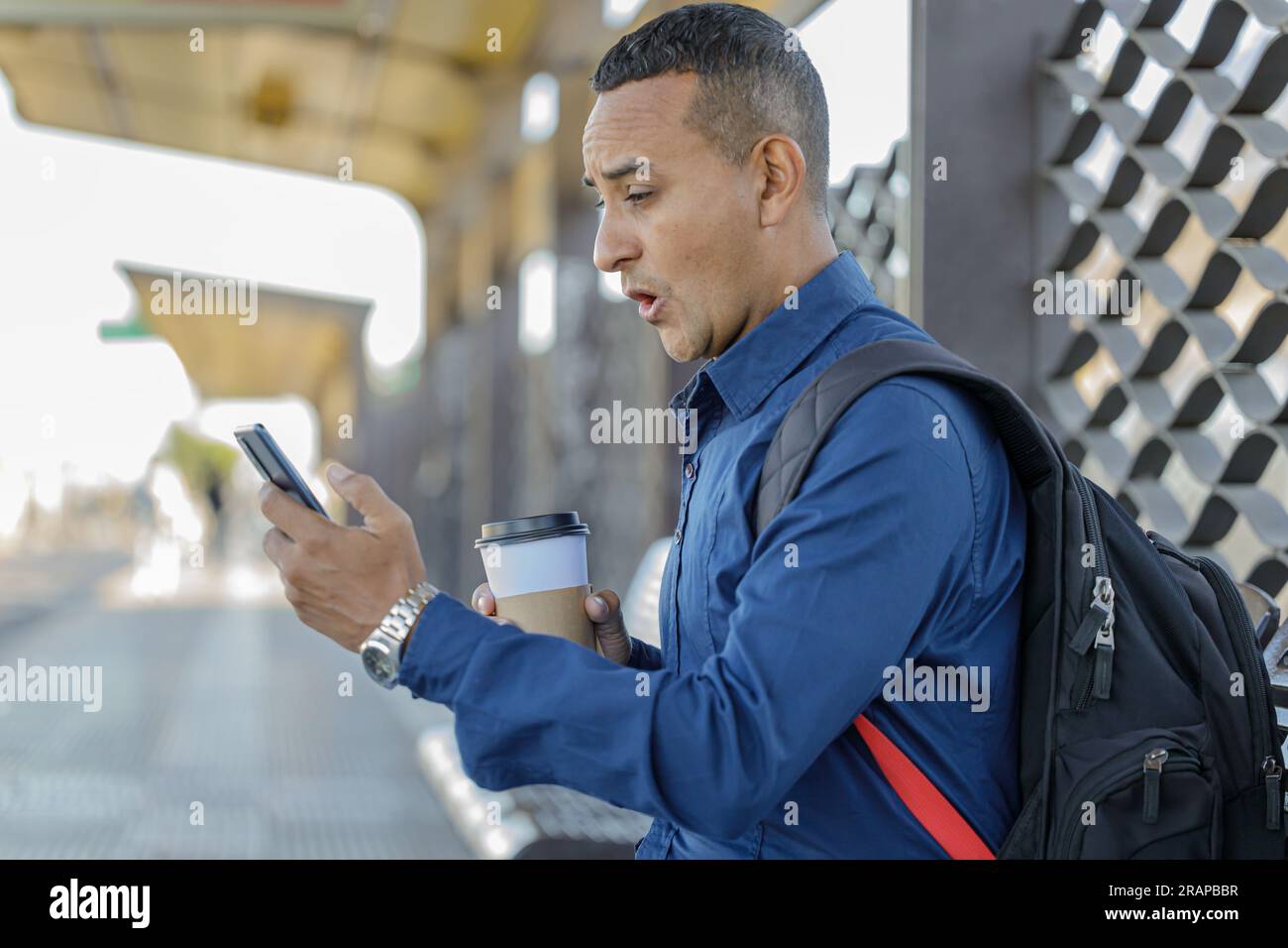 Young latin man looking at his mobile phone with a surprised face a paper cup with coffee in his hand. Stock Photo