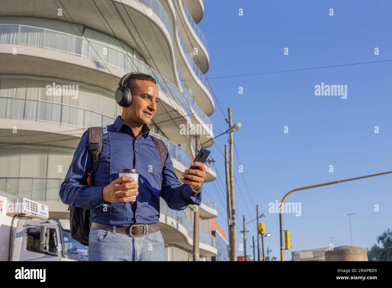 Young latin man with headphones looking at his mobile phone and holding a paper cup with coffee in his hand with copy space. Stock Photo