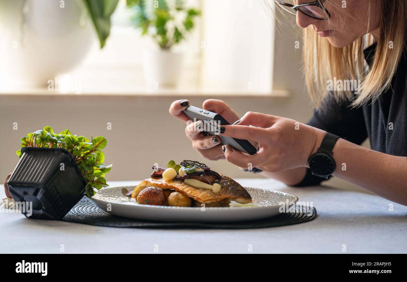 A woman taking photo with smart phone of roasted branzino with mushrooms and butter potatoes. Posting and sharing food pictures on social media. Stock Photo
