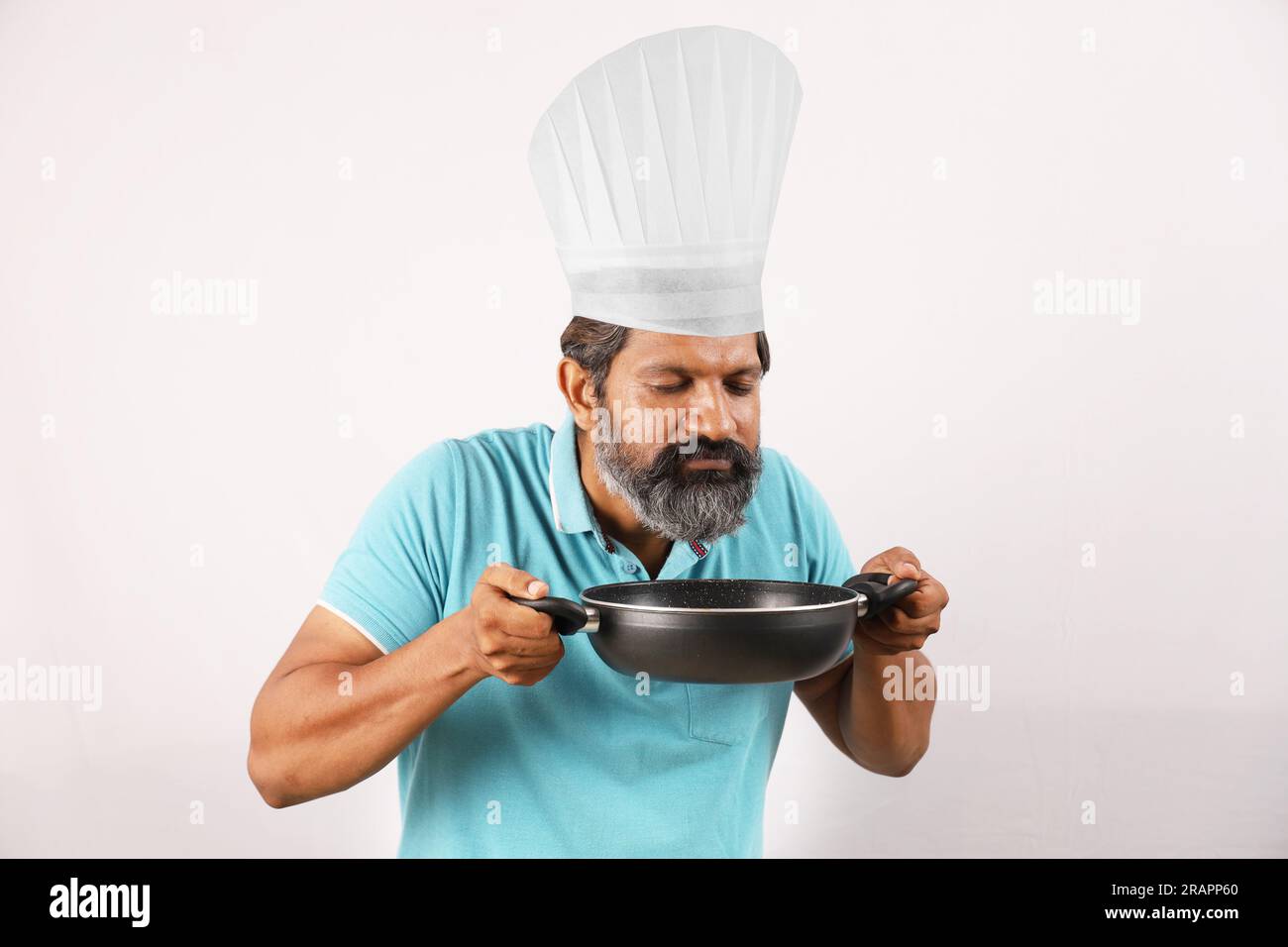 Portrait of Indian bearded chef man wearing t-shirt and chef cap. Happy smiling expressions white background. Holding spatula and frying spoon in hand Stock Photo