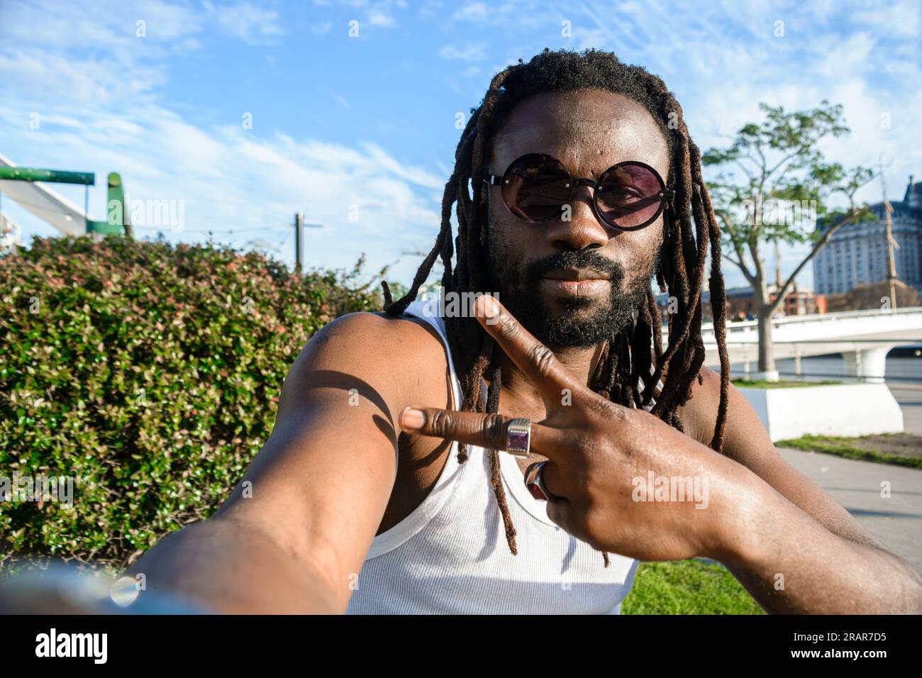 Selfie portrait of young African man with dreadlocks, glasses and beard sitting looking at the camera making v sign with hand and smiling with good at Stock Photo
