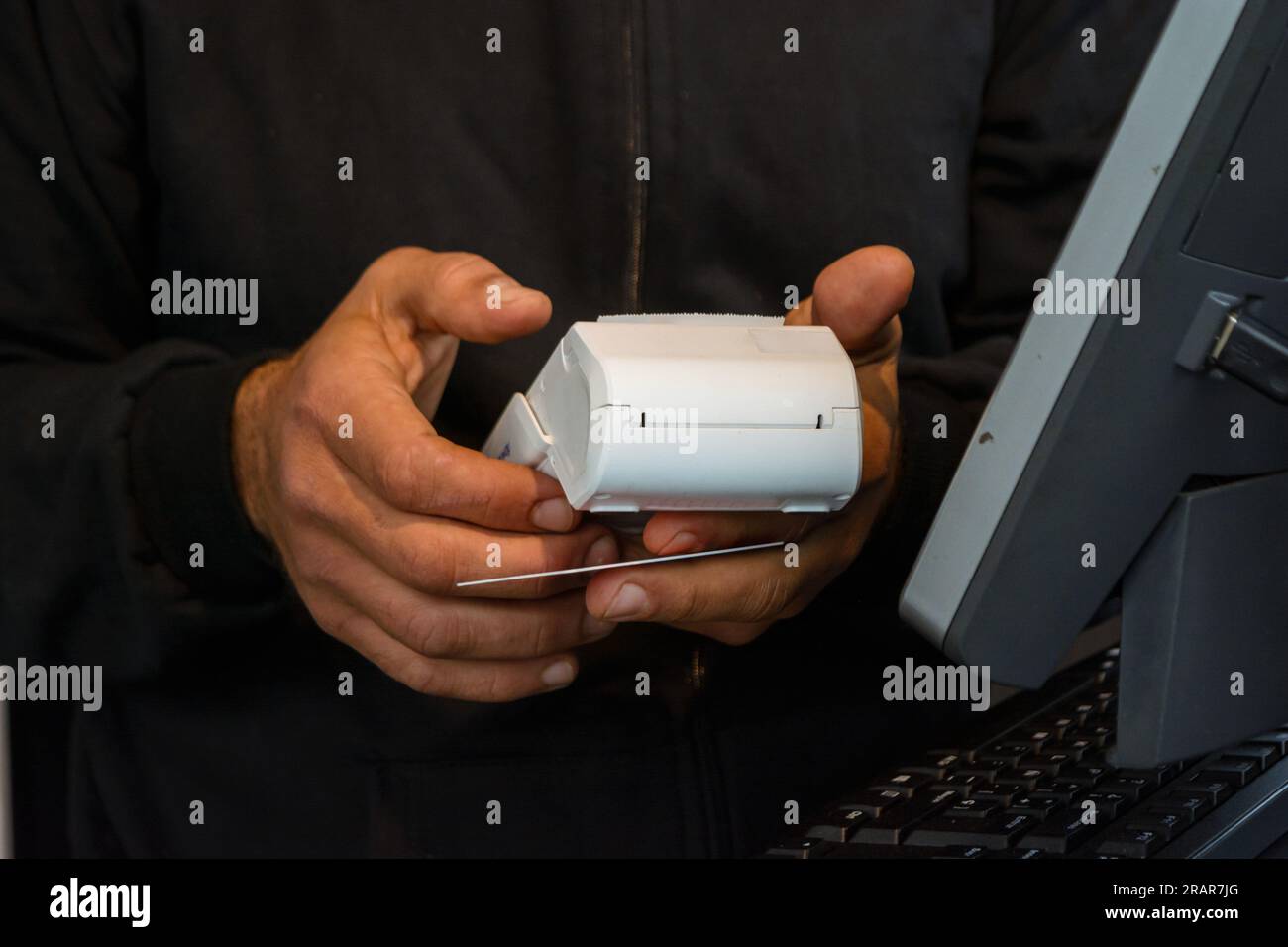 Male hands of unrecognizable waiter dressed in black, holding electronic debit card payment point, cashing out next to computer monitor. Stock Photo