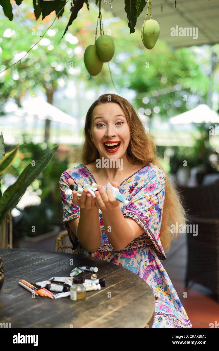 Portrait of beautiful blonde woman artist and illustrator  sitting by the table and happily holding her acrylic paints Stock Photo