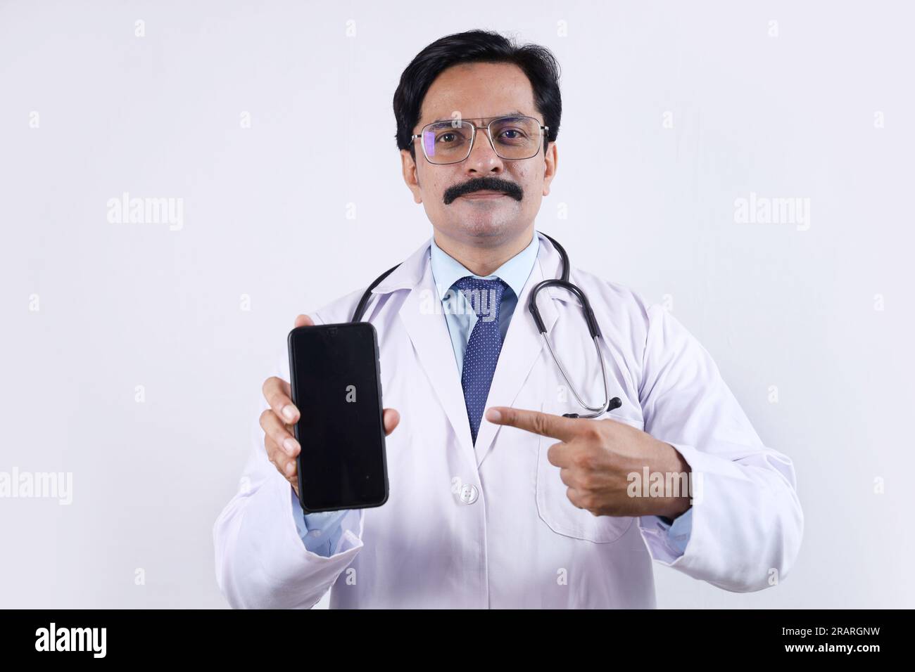 Beautiful portrait of happy Indian doctor in uniform wearing stethoscope and specs showing phone screen in hand. Showing mobile app. Stock Photo