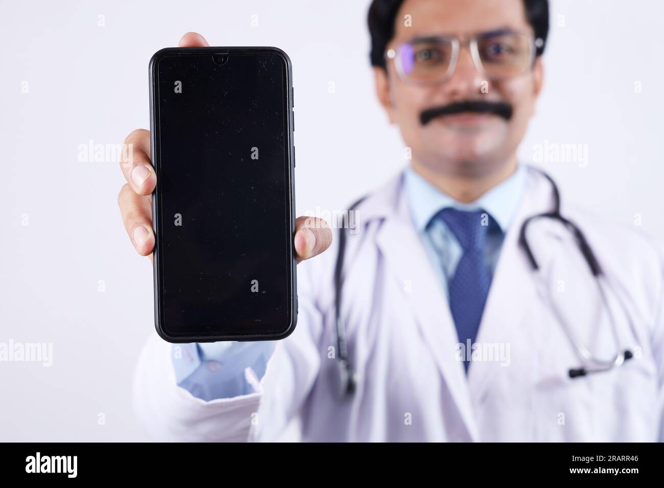 Beautiful portrait of happy Indian doctor in uniform wearing stethoscope and specs showing phone screen in hand. Showing mobile app. Stock Photo