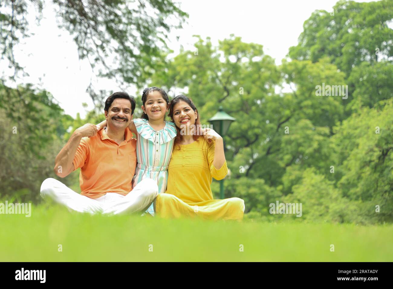 Happy Indian family with a girl child sitting together on grass and enjoying their family time early morning. The family is looking at the camera. Stock Photo