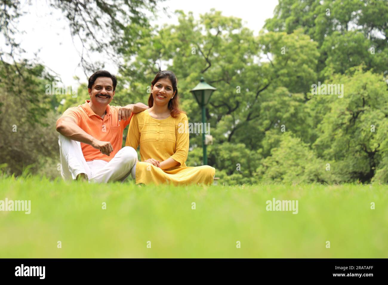 Young happy couple leaning together on grass in a public park early morning in positive atmosphere. Green, tranquil and serene environment. Stock Photo