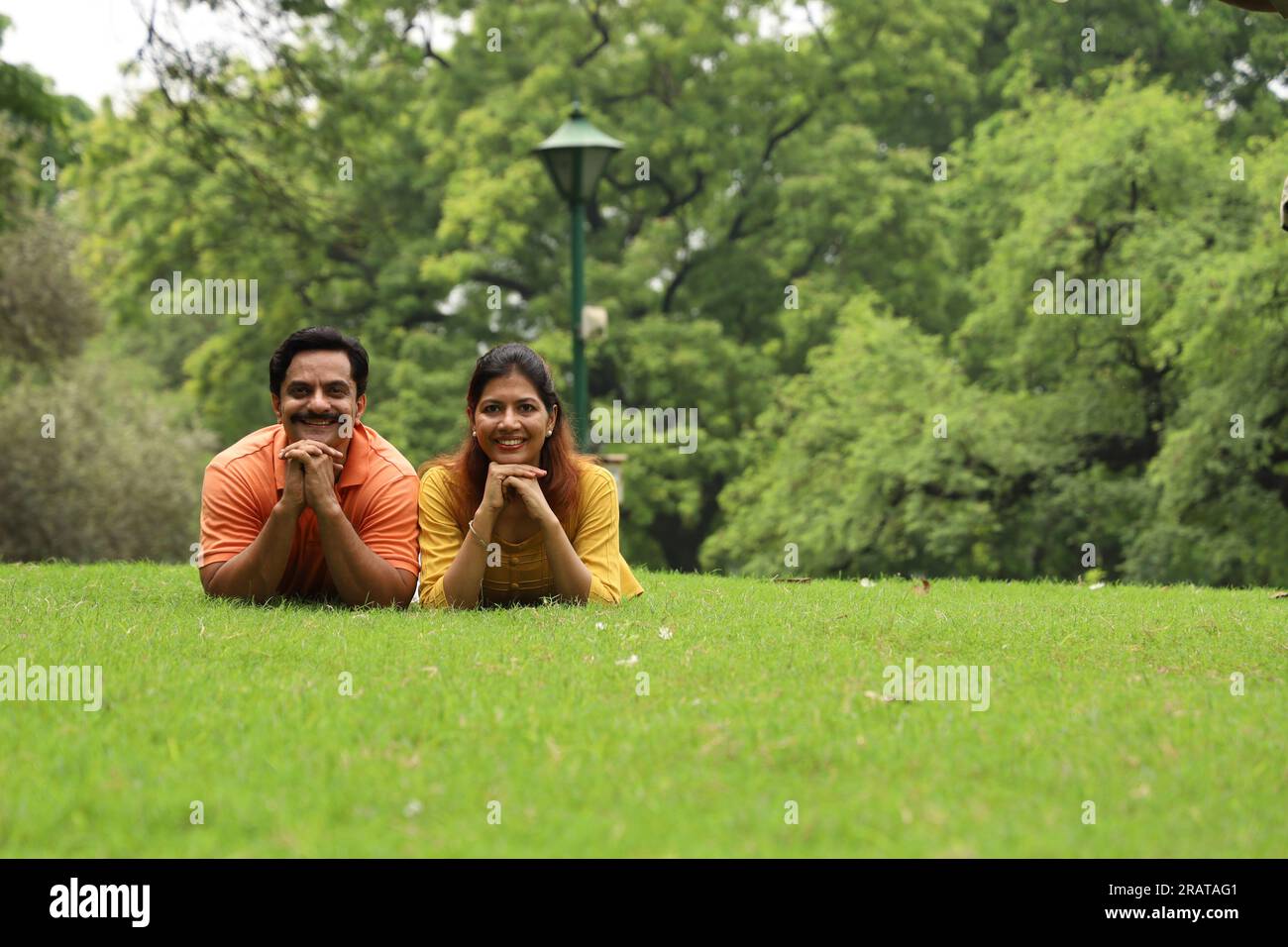 Young happy couple leaning together on grass in a public park early morning in positive atmosphere. Green, tranquil and serene environment. Stock Photo