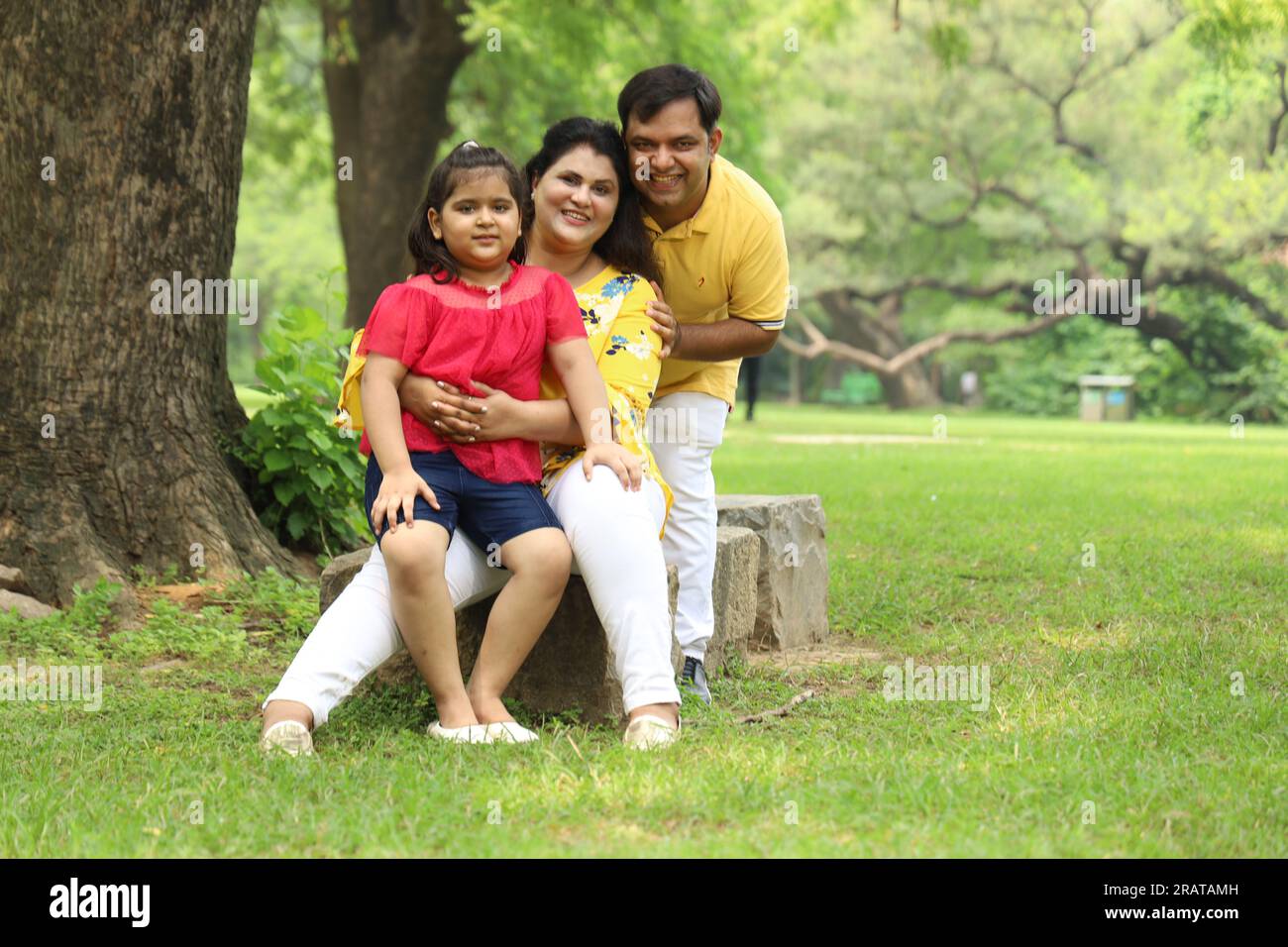 Happy Indian family with a girl child sitting together on grass and enjoying their family time early morning. The family is looking at the camera. Stock Photo