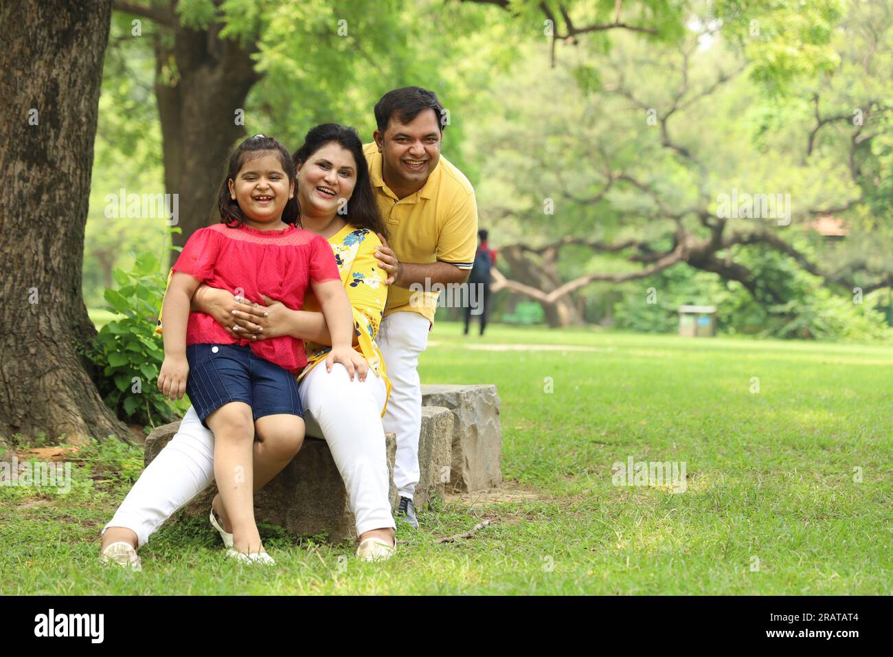 Happy Indian family with a girl child sitting together on grass and enjoying their family time early morning. The family is looking at the camera. Stock Photo