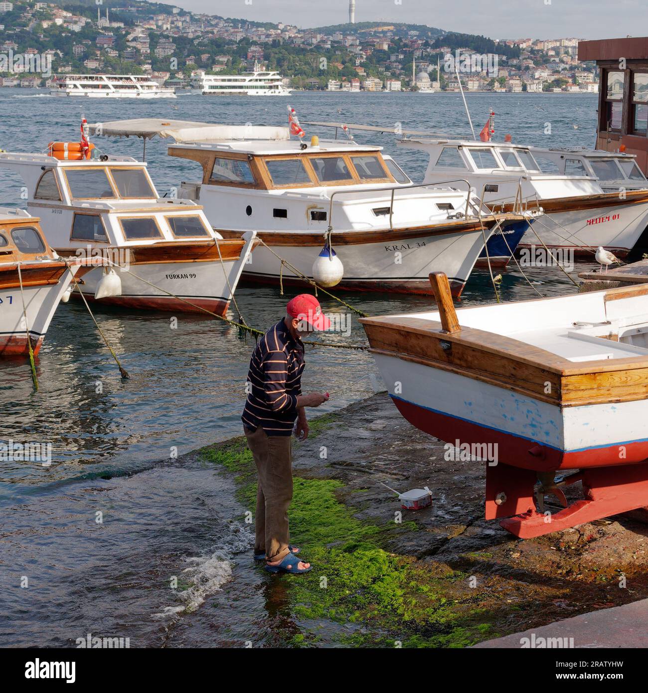 Man paints a wooden boat on a summers evening by the Bosporus shoreline in Istanbul. Passenger ferries and the Asian side in the background. Turkey Stock Photo