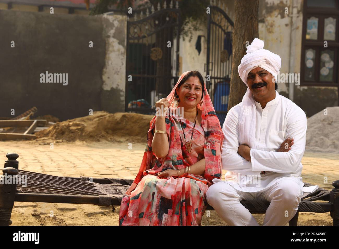 Happy Indian Rural family in village. Husband, wife sitting on cot outside their home in front yard. man in kurta pajamas and wife wearing saree, sari Stock Photo