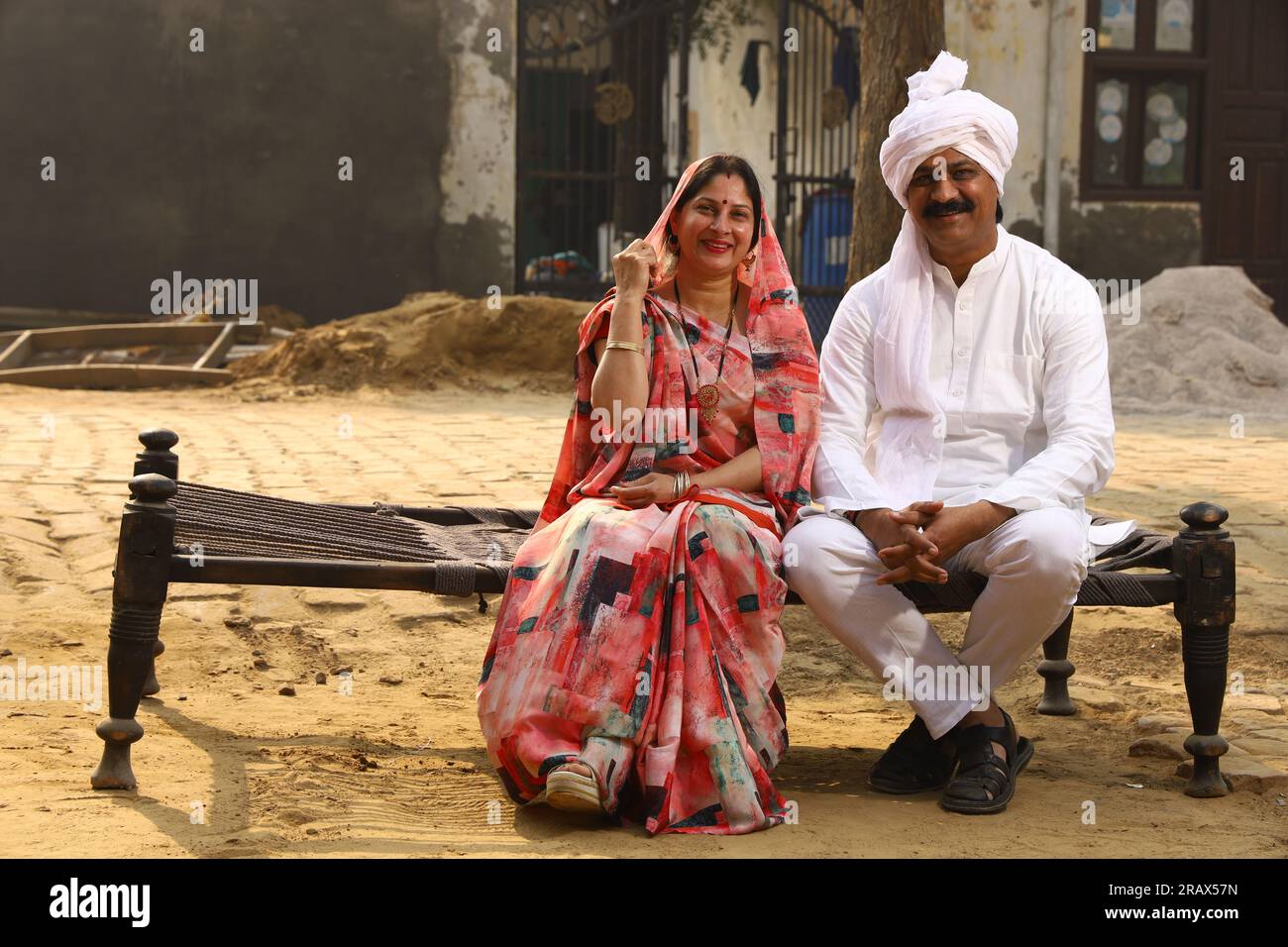 Happy Indian Rural family in village. Husband, wife sitting on cot outside their home in front yard. man in kurta pajamas and wife wearing saree, sari Stock Photo