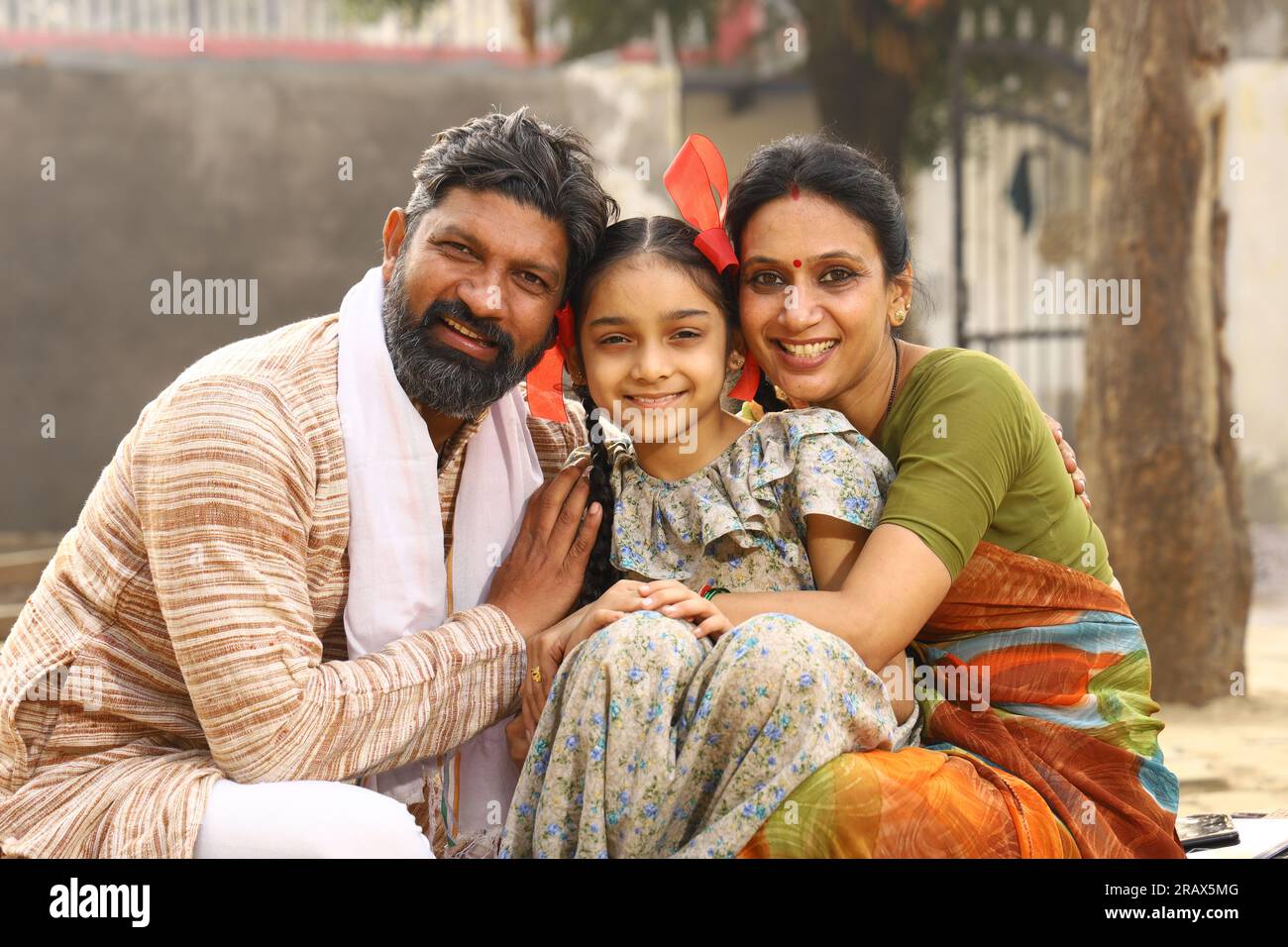 Happy rural Indian family sitting together outside their cottage in day time smiling while looking at the camera. Girl Child Concept. Stock Photo