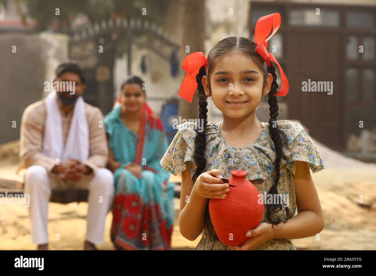 Happy rural Indian family sitting together, daughter is holding a piggy bank in her hand, putting a coin in it, showing the concept of saving money Stock Photo