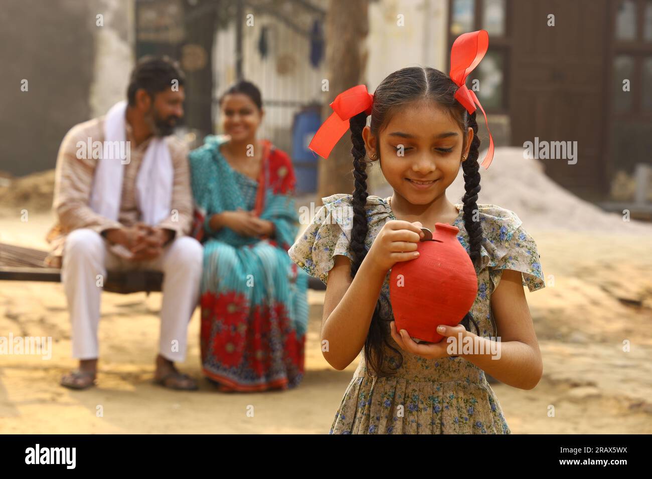 Happy rural Indian family sitting together, daughter is holding a piggy bank in her hand, putting a coin in it, showing the concept of saving money Stock Photo