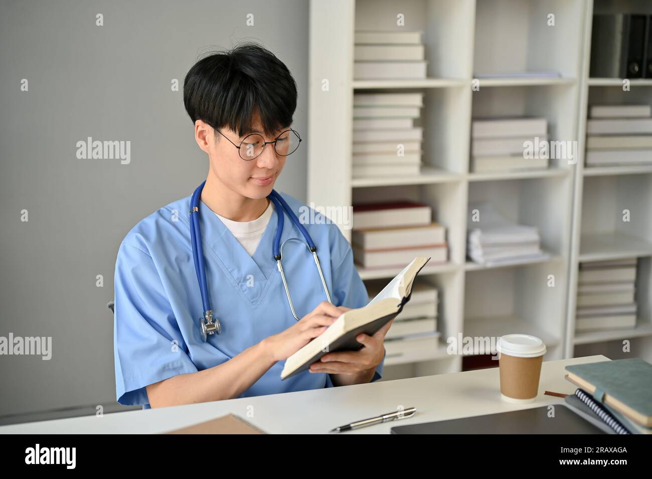 A smart and focused young Asian male medical student in scrubs reading ...