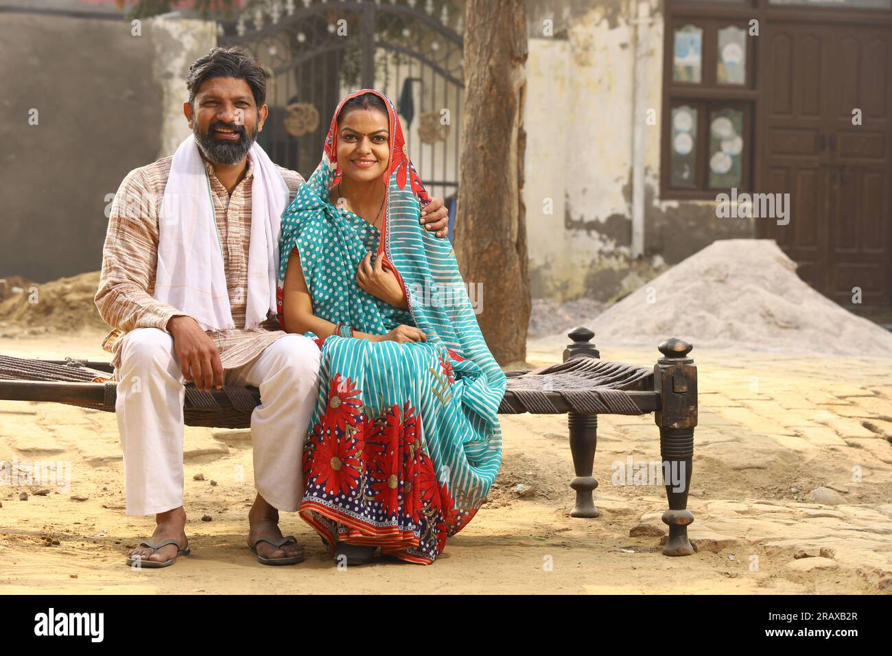 Happy Indian Rural family in village. Husband and wife sitting on cot outside their home front yard. man in kurta pajamas and wife in beautiful saree. Stock Photo