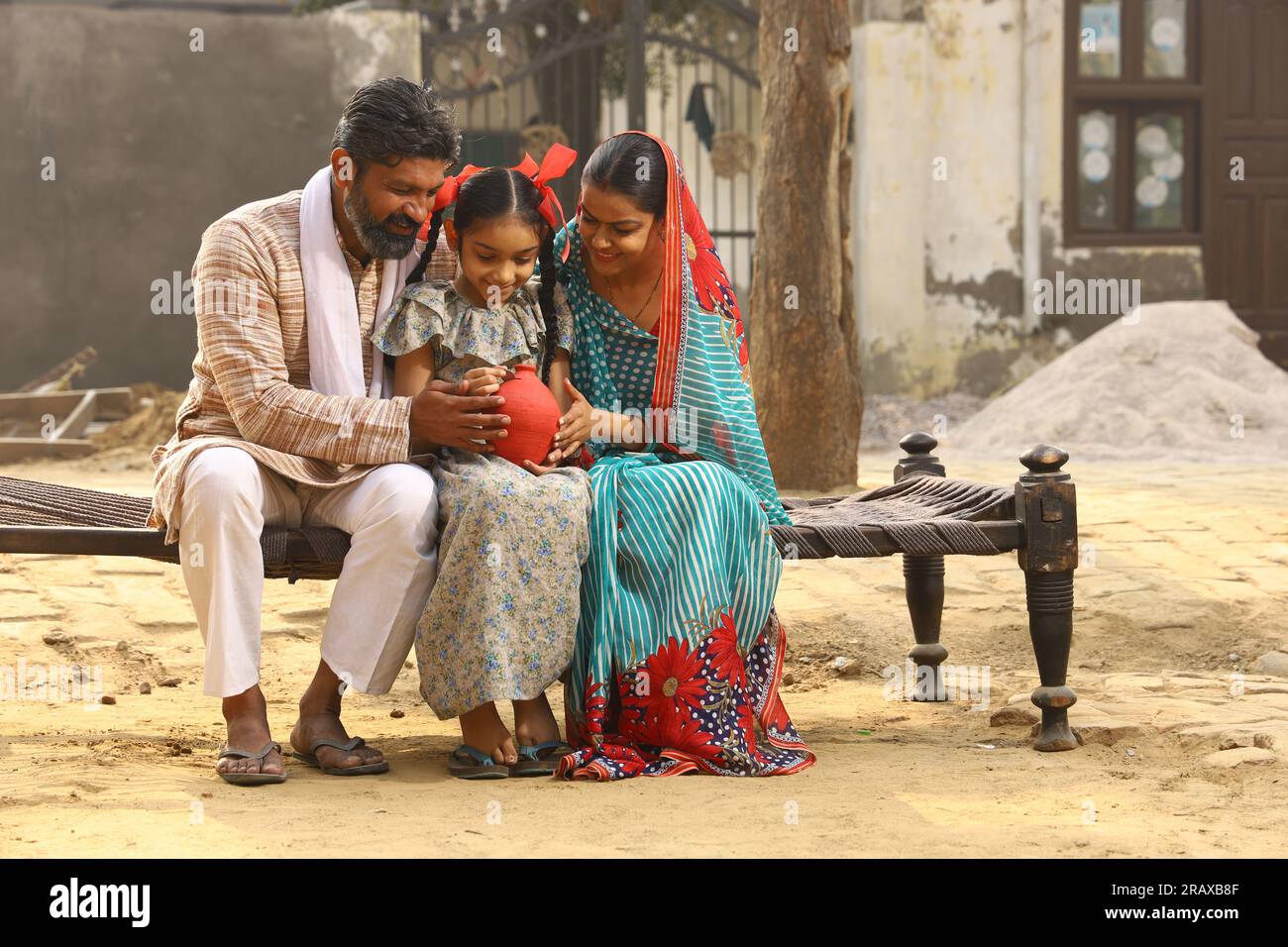 Happy rural Indian family sitting together, daughter is holding a piggy bank in her hand, putting a coin in it, showing the concept of saving money Stock Photo