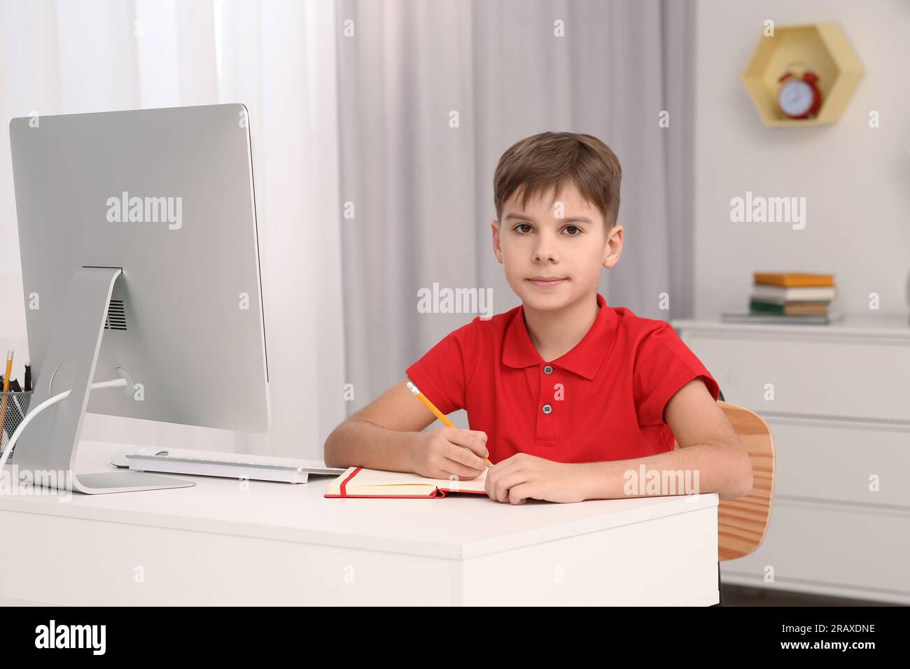 Boy writing in notepad while using computer at desk in room. Home workplace Stock Photo