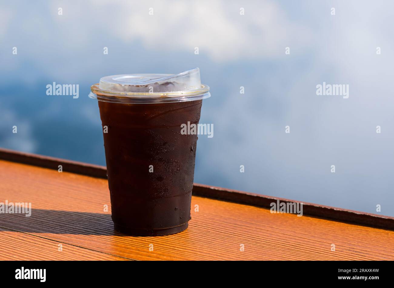 A glass of iced coffee with iced cube puts on table with sky and cloud