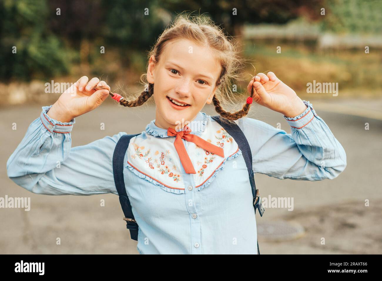 Joyful little schoolgirl looking straight at camera, holding pigtais, wearing vintage uniform and backpack Stock Photo