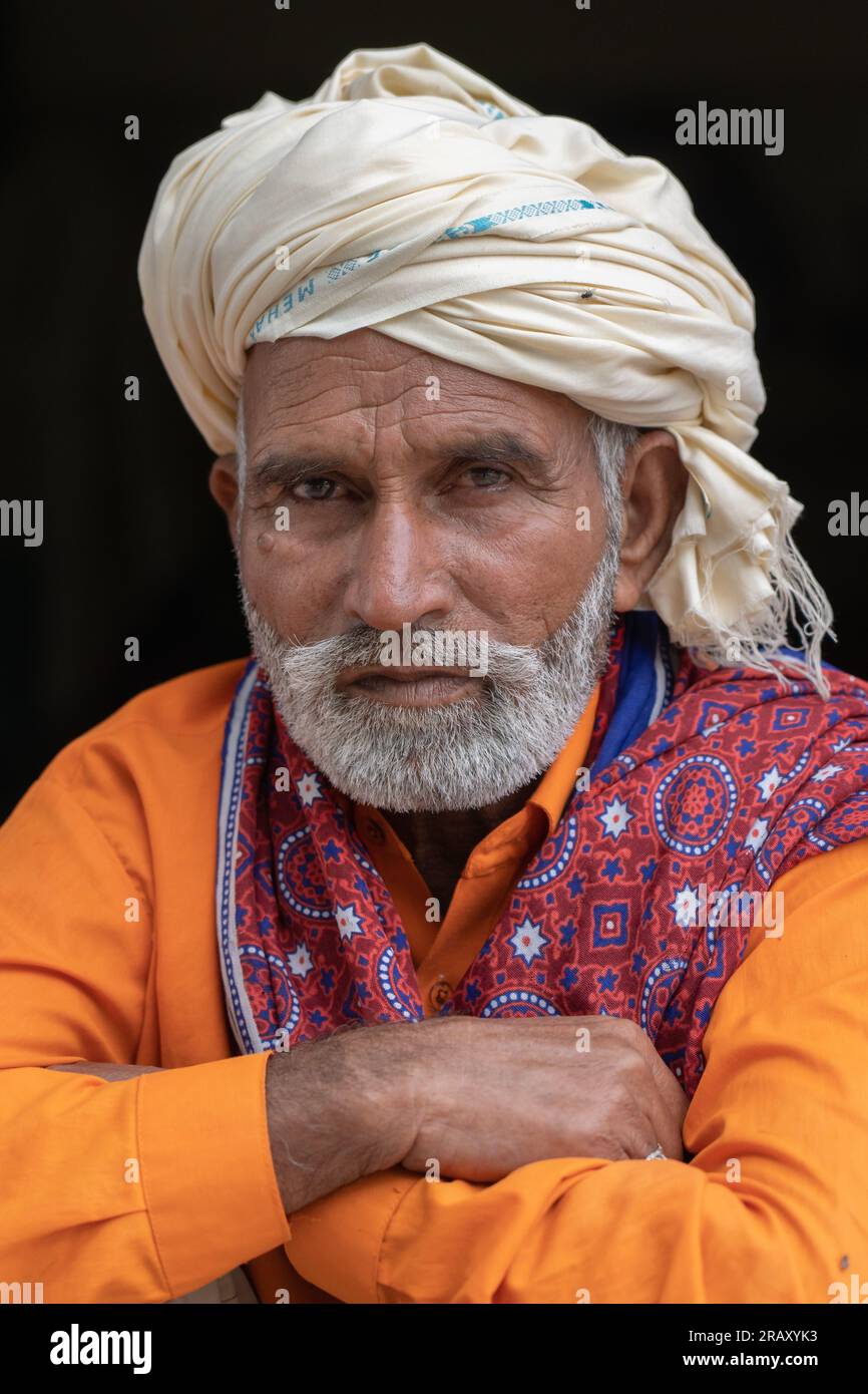 Handsome middle aged Pakistani man with beard and orange shirt wearing traditional white turban and ajrak shawl on black background Stock Photo