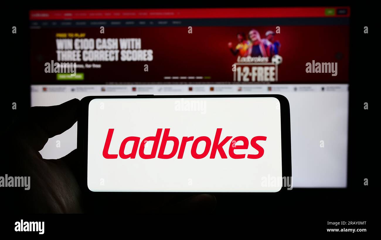 Person holding mobile phone with logo of company LC International Limited (Ladbrokes) on screen in front of web page. Focus on phone display. Stock Photo