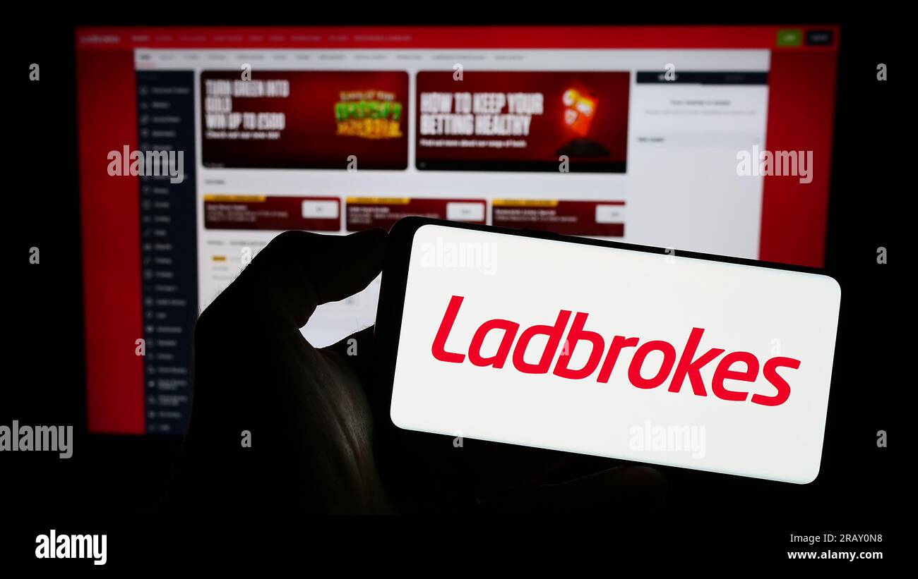 Person holding cellphone with logo of company LC International Limited (Ladbrokes) on screen in front of webpage. Focus on phone display. Stock Photo