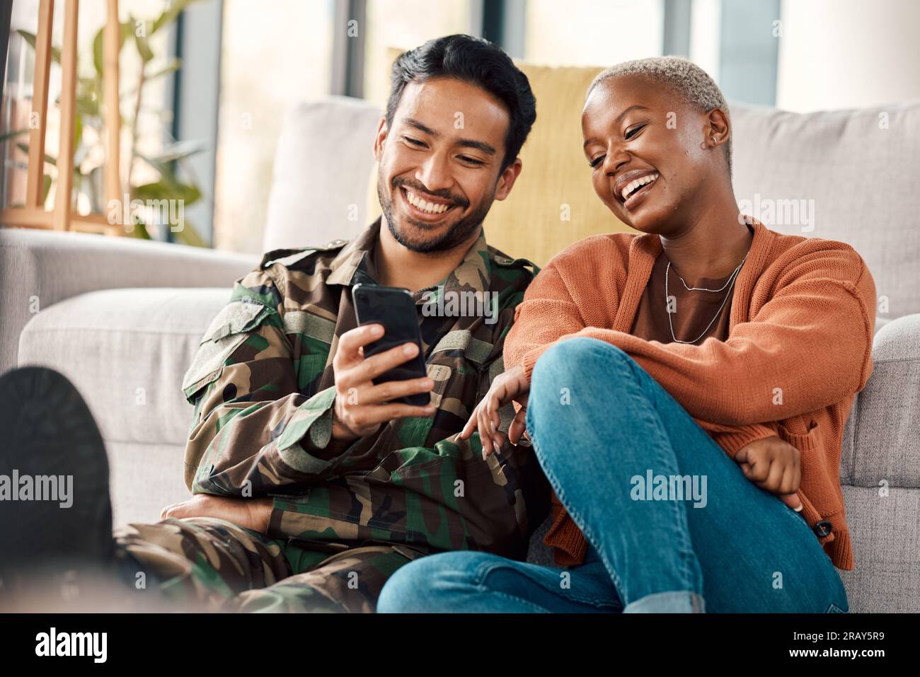 Army man, interracial couple and return with phone meme at home with laugh and video streaming. Lounge, young people and mobile scroll together in a Stock Photo