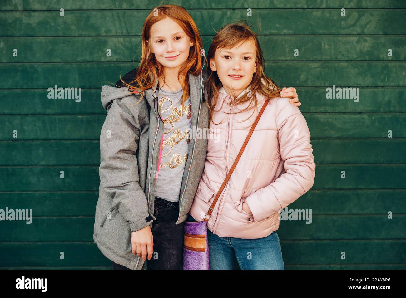 Outdoor portrait of two cute little teen girls wearing warm jackets, posing against green background, fashion for children Stock Photo