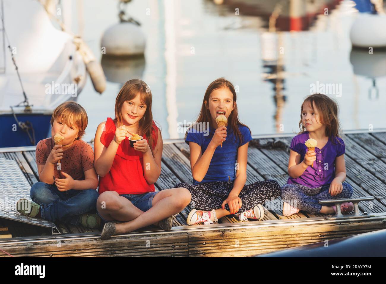 Group of four kids, 3 girls and one boy, eating ice cream outdoors, resting on a pier by the lake Stock Photo