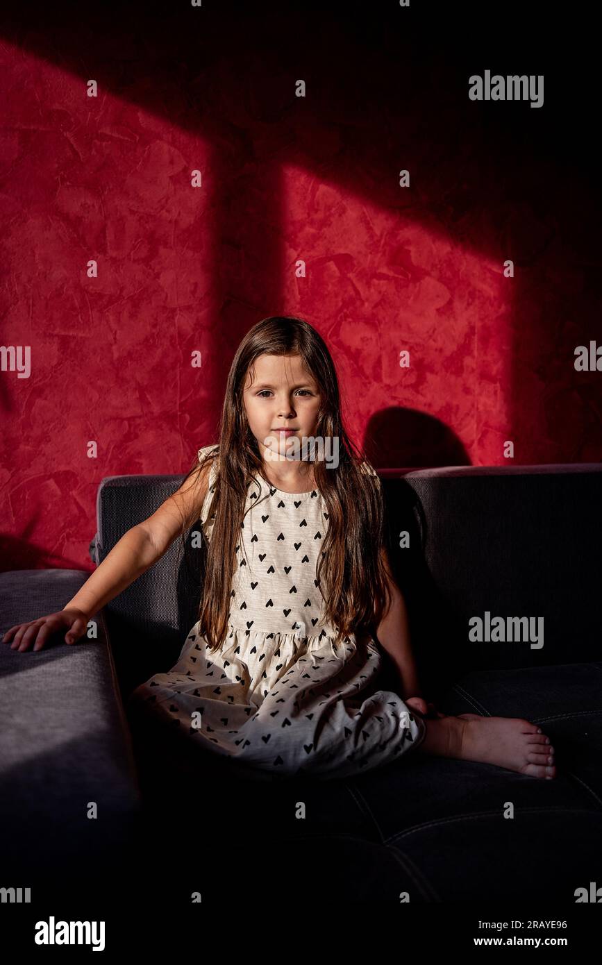 Portrait of little girl with long hair sits on sofa against red ...