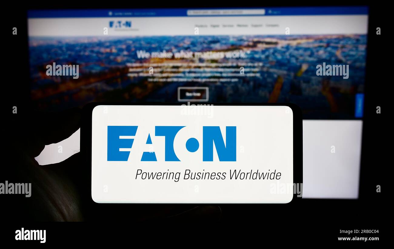 Person holding cellphone with logo of power management company Eaton Corporation plc on screen in front of webpage. Focus on phone display. Stock Photo