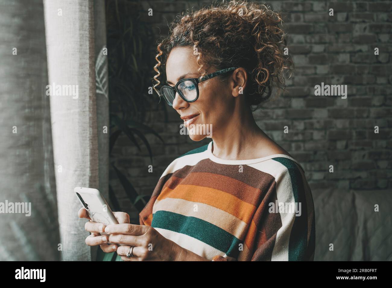 One happy woman indoor smiling at the phone and using app to chat or message people. Dating and lonely lifestyle female people with technology and internet connection. Young lady writing on cellular Stock Photo