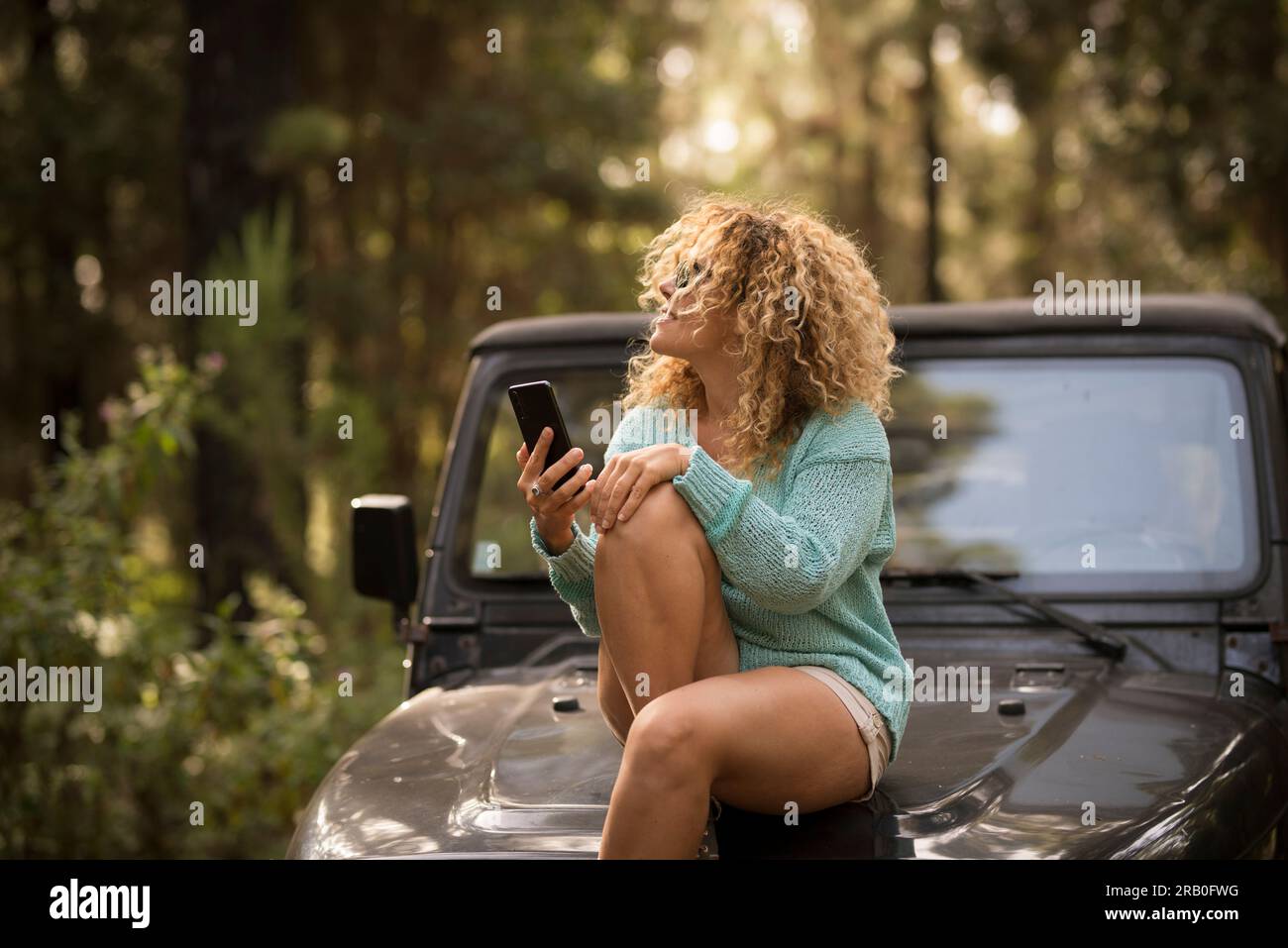 Young tourist lady enjoy car travel destination in the natural park sitting on the vehicle and using mobile phone. Traveler and adventure lifestyle. Weekend outdoor leisure activity alone. One woman Stock Photo