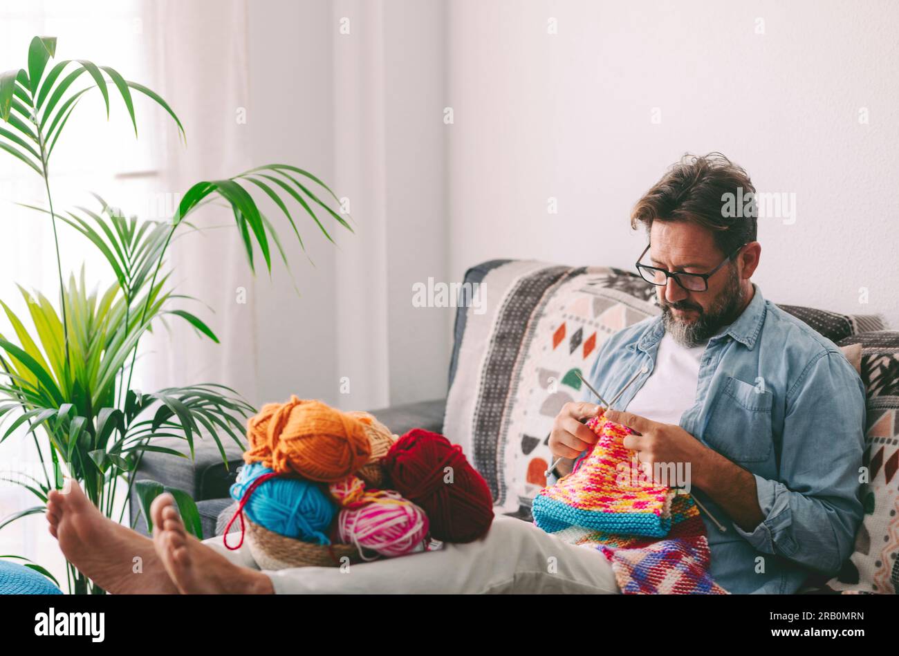 One man at home sitting on the sofa in relaxation indoor leisure activity alone doing knitting work with colorful wool and needles. Learning new things job in life. Relax lifestyle male people at home Stock Photo