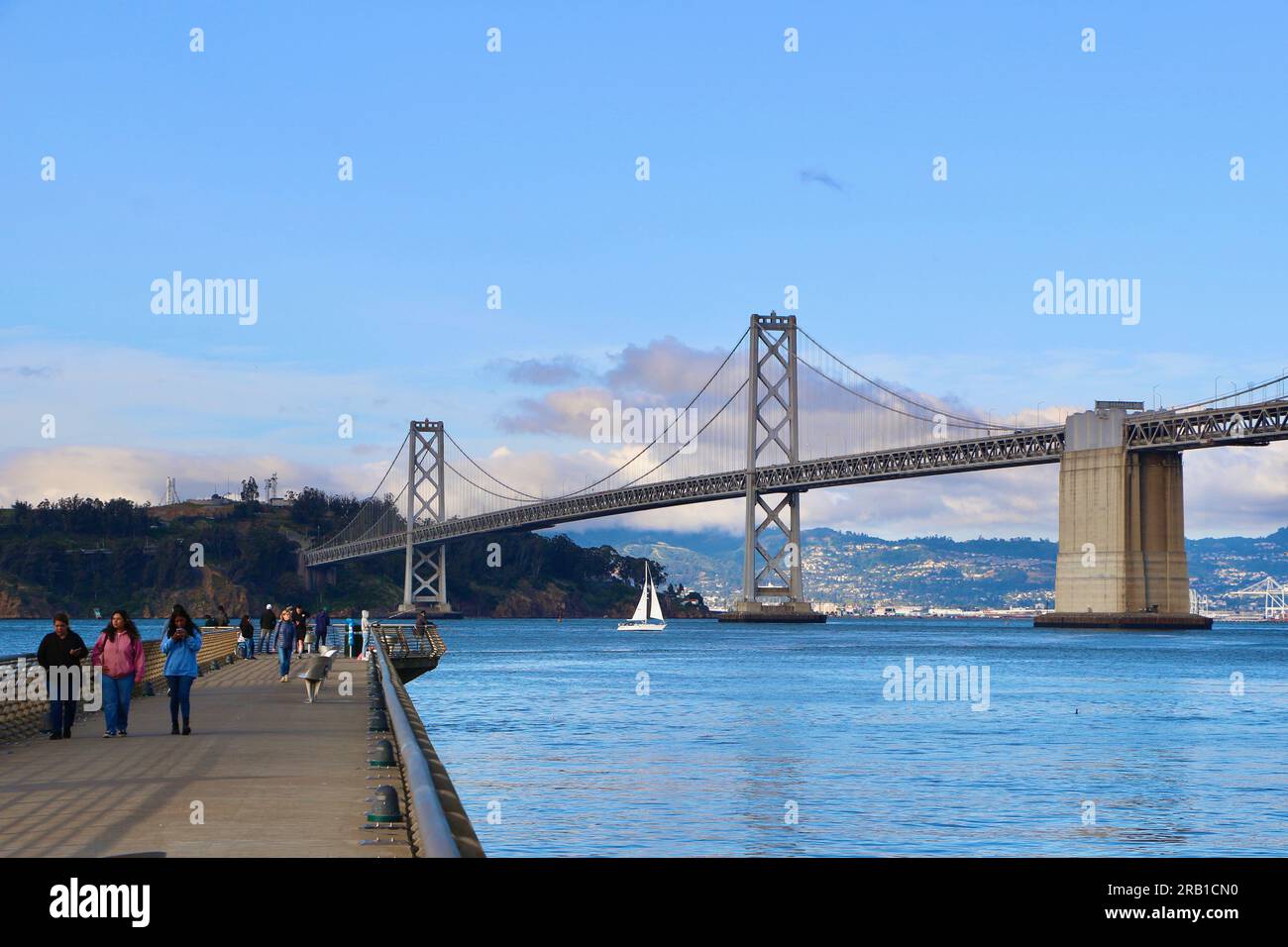 Tourists on Pier 14 with a landscape view of the bay and Bay Bridge with a passing yacht under full sail The Embarcadero San Francisco California USA Stock Photo