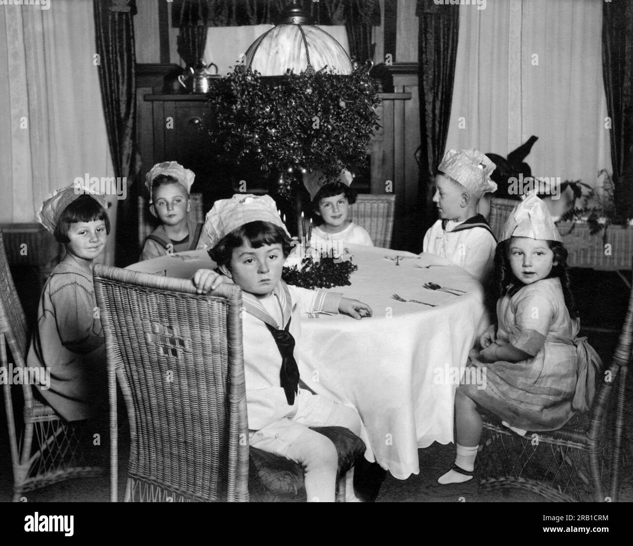 United States:  c. 1910. Six young children wearing party hats sitting around a table anxiously awaiting... Stock Photo
