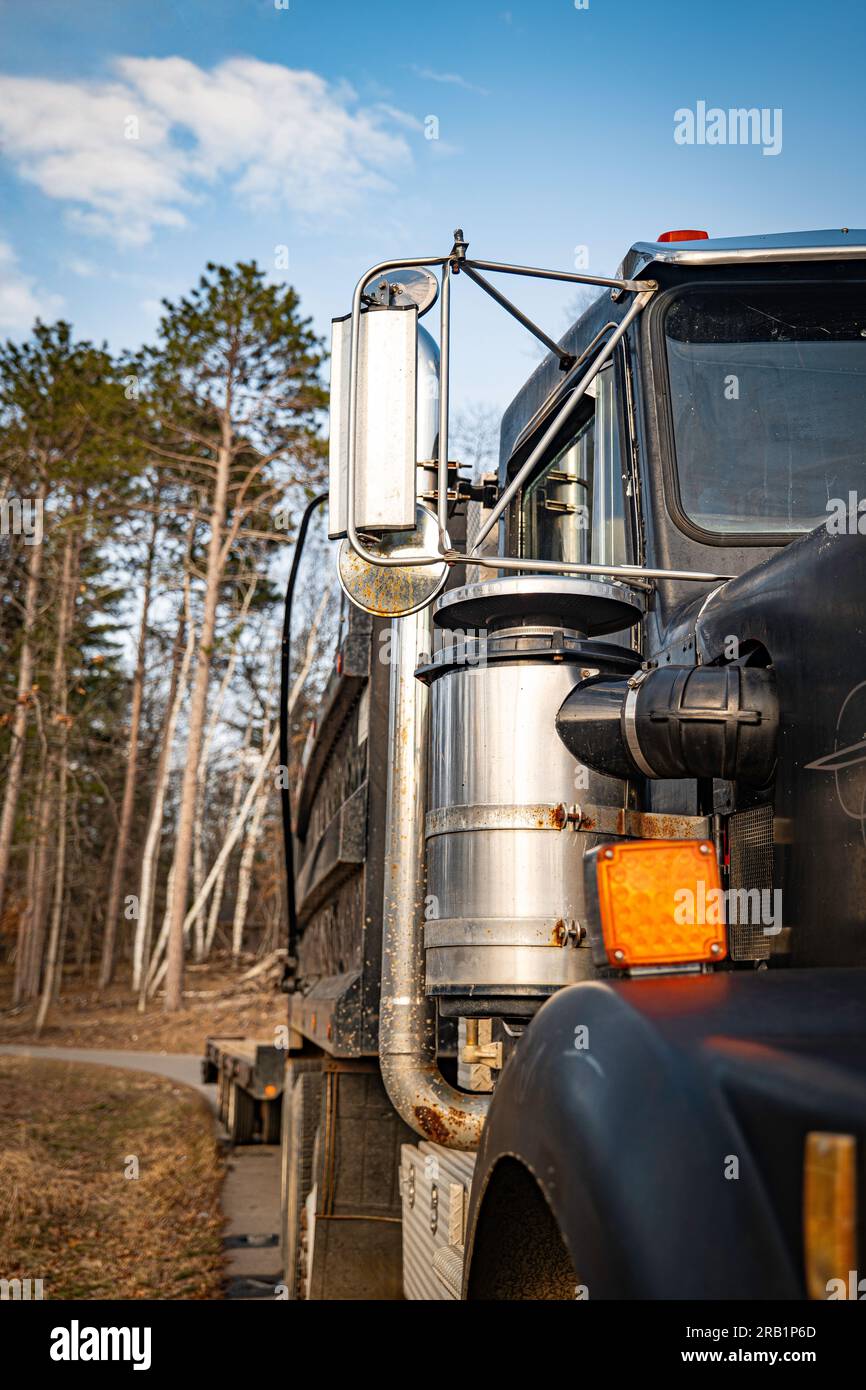 Side view of a black dump truck and trailer parked on a paved road on a sunny afternoon, with trees in distance, and mirror, air intake shroud, exhaus Stock Photo