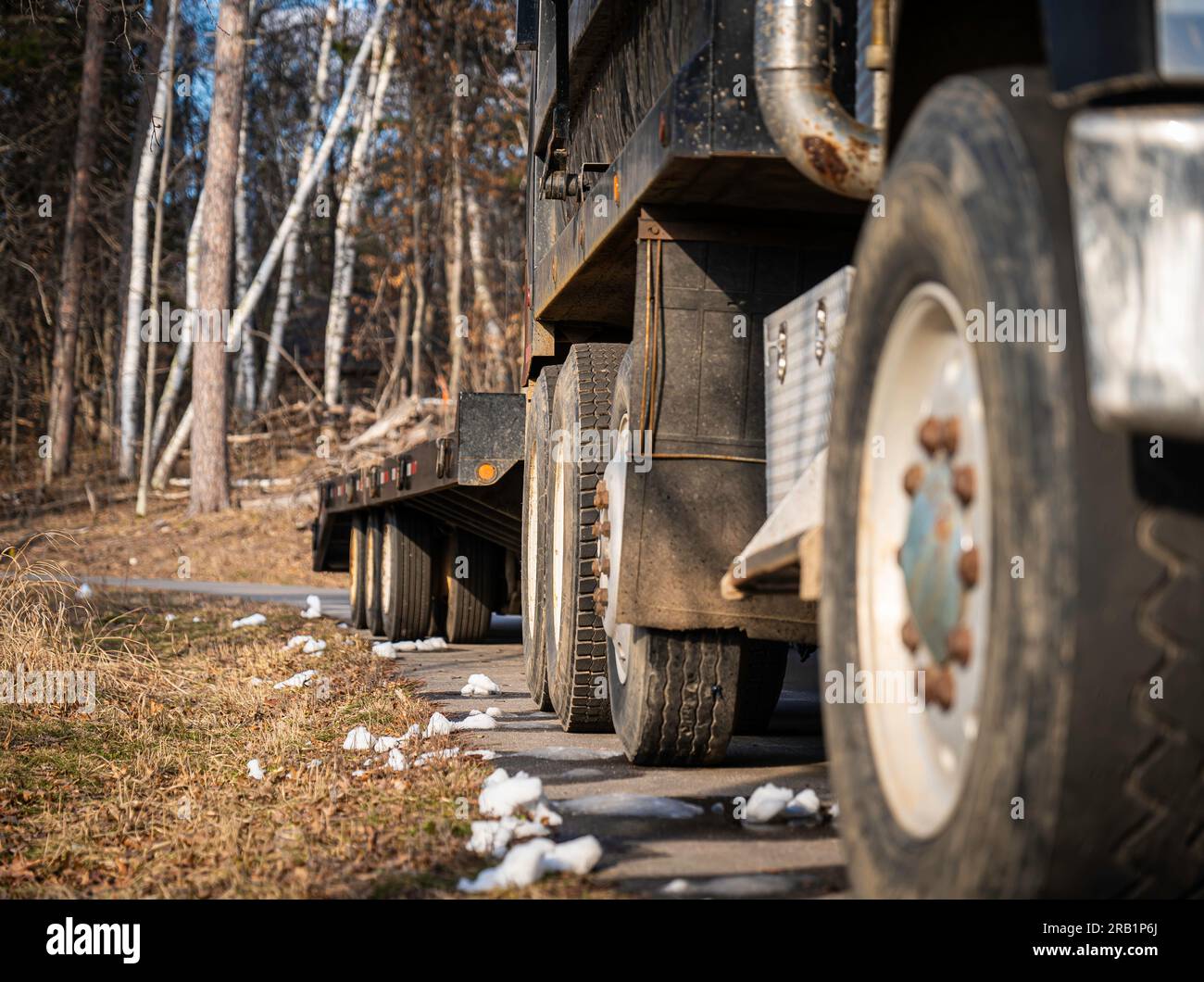 Tires and wheels and right side of a truck and trailer parked on a paved road on a sunny afternoon, with melting chunks of snow on the ground at job s Stock Photo