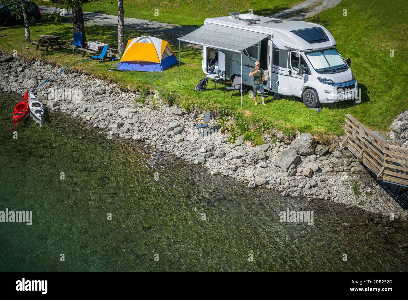 Aerial View of Motorhome Traveler Enjoying Coffee in Front of His RV After Setting Up a Campsite by the River. Camping and Traveling Theme. Stock Photo