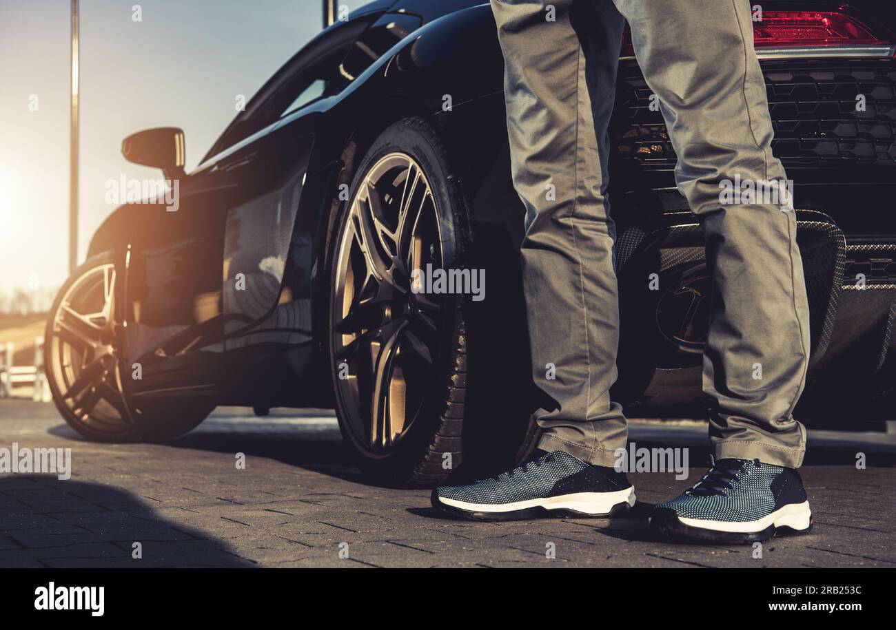 Closeup of Male's Legs in Sports Shoes Next to Modern Sport Car. Luxury Vehicle Owner. Sunset Light in the Background. Successful Lifestyle Theme. Stock Photo