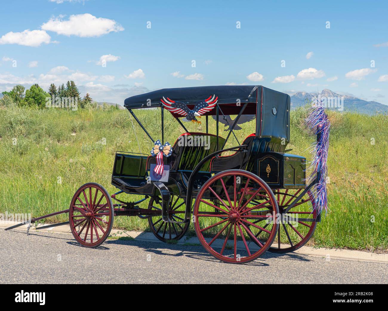 Surrey carriage over 100 years old near field & mountain background is parked & decorated in USA red, white, & blue. See 2 bench seats & red wheels. Stock Photo