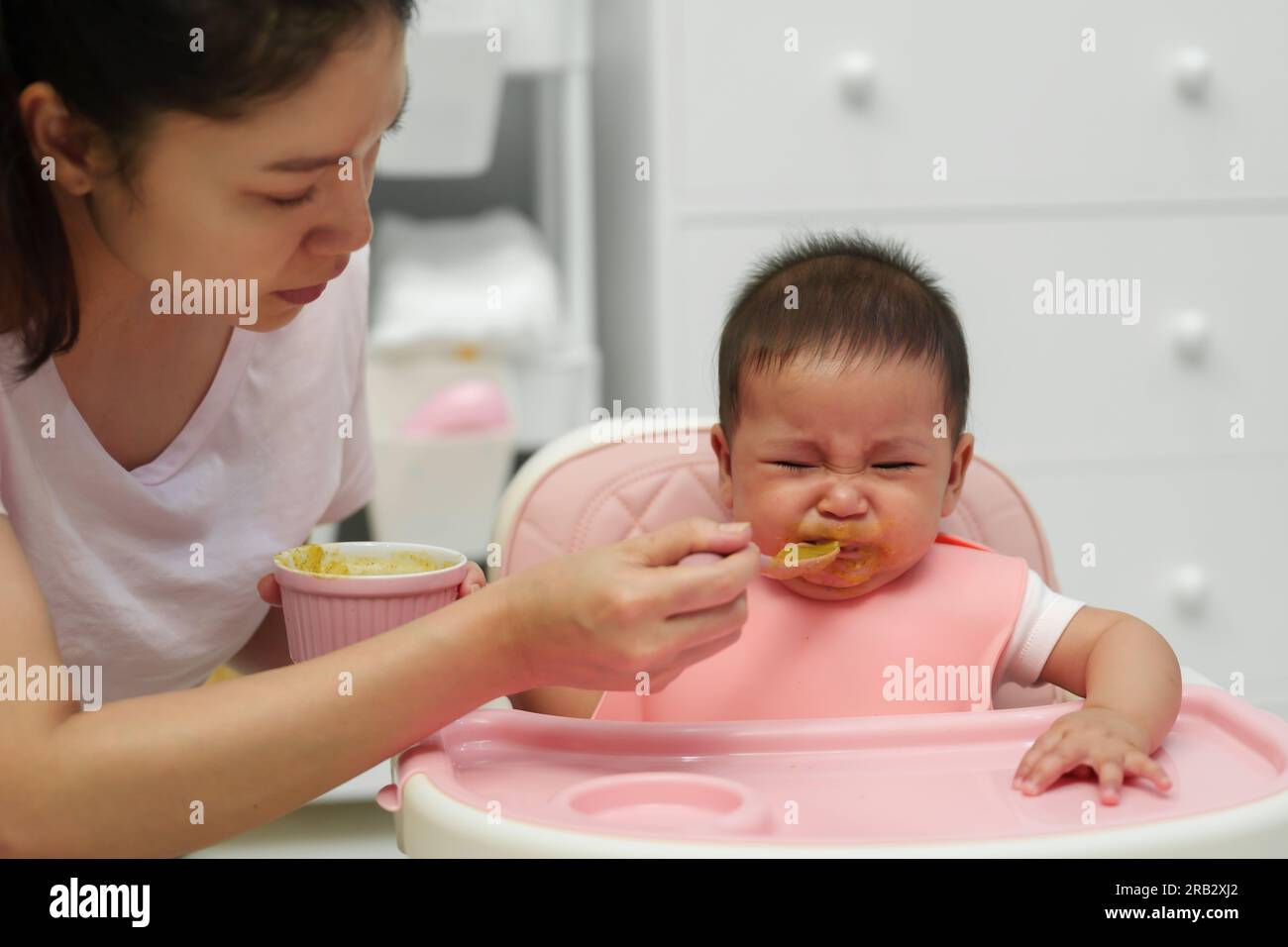 mother feeding food to her crying infant baby with a spoon, refuse to eat Stock Photo