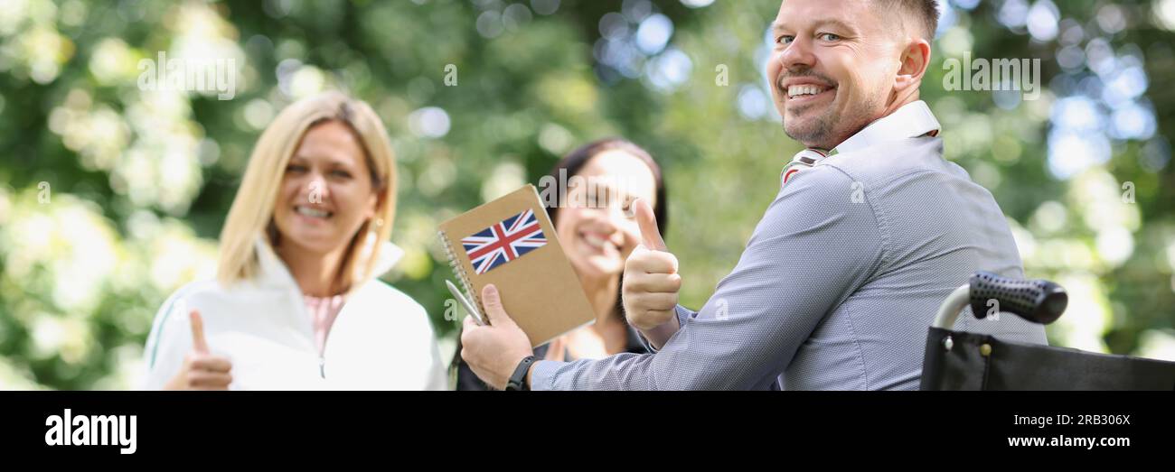 Smiling disabled man in wheelchair studying english with friends and showing thumbup sign Stock