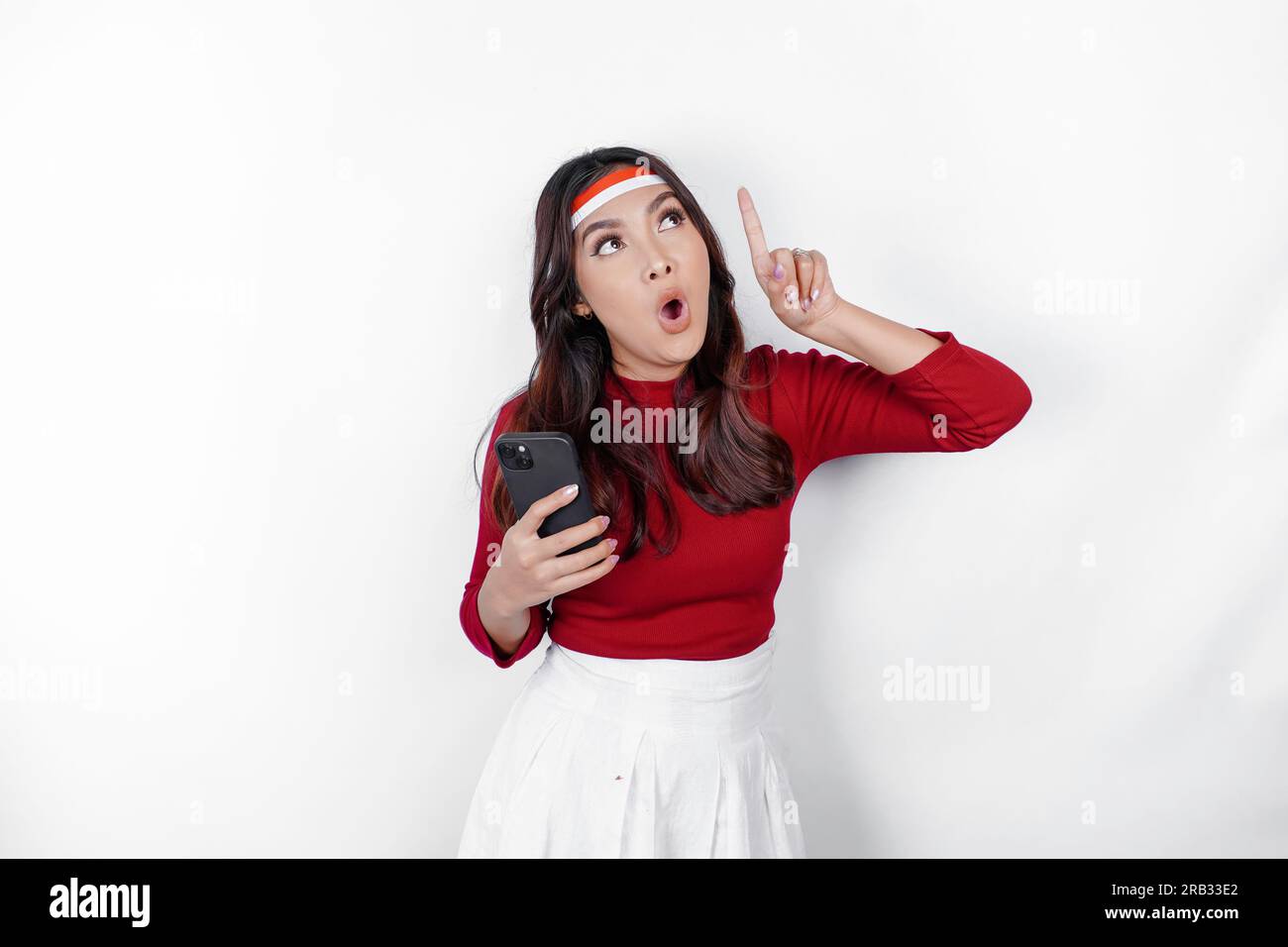 A portrait of a shocked Asian woman wearing headband, holding her phone, and pointing copy space on top of her, isolated by white background. Indonesi Stock Photo